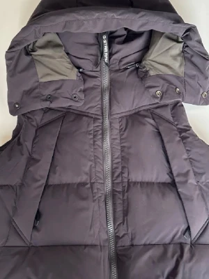 Svart dunväst från G-Star RAW - Snygg svart dunväst från G-Star RAW med hög krage och dragkedja framtill. Västen har en avtagbar huva med tryckknappar och två sidofickor. Insidan är fodrad i olivgrönt tyg och materialet är quiltat för extra värme. Perfekt för kalla dagar och streetwear-stil. Västen är helt oanvänd med original tags kvar. Original pris 2199kr