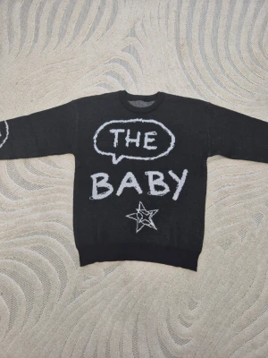 Svart stickad tröja med text och stjärna - Cool svart stickad tröja med vit text 'THE BABY' och en stjärna framtill. Tröjan har rund halsringning och långa ärmar, samt vita detaljer på ärmarna. Perfekt för dig som gillar streetstyle och unika plagg.
