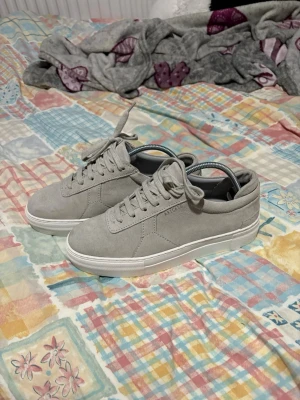 Gråa sneakers från Arigato i mocka - Stilrena gråa sneakers från Axel Arigato i mjuk mocka med vita sulor och klassisk snörning. Diskret logga på sidan och låg profil som passar till det mesta.