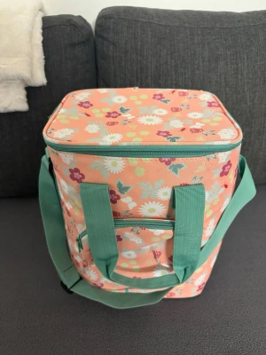 Blommig kylväska med mintgröna detaljer - Säljer en söt kylväska i persikofärg med blommigt mönster och mintgröna handtag samt axelrem. Väskan har en fyrkantig form, dragkedja och en liten ficka framtill. Perfekt för picknick eller utflykt. Materialet är tålig textil.