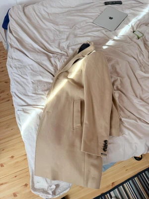 Beige klassisk ullkappa - Stilren beige kappa i ullblandning med rak passform och snygga detaljer som knappar vid ärmslut och fickor framtill. Perfekt för dig som vill ha en tidlös och trendig look. Passar lika bra till jeans som till kostymbyxor.