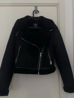 Svart skinnjacka från Pull&Bear - Snygg svart skinnjacka från Pull&Bear med silvriga dragkedjor och mjukt foder. Jackan har en klassisk bikerstil med bred krage och två snedställda fickor med dragkedja. Jackan passar perfekt vädret just nu!!