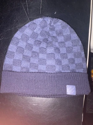 Blå Louis Vuitton mössa i ullmix - Säljer en snygg blå mössa från Louis Vuitton med klassiskt schackrutigt mönster och vikt kant. Märkeslapp framtill och mjuk ullblandning som håller dig varm. Perfekt accessoar för dig som vill ha något exklusivt och stilrent.