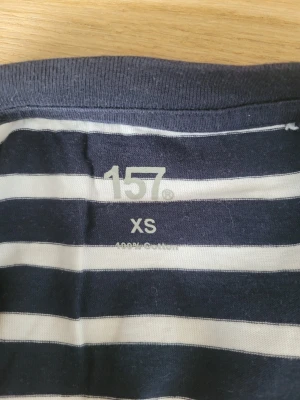 Randig långärmad tröja 157 XS - Mörkblå och vit långärmad tröja från 157 i storlek XS. Tröjan har klassiska horisontella ränder och är tillverkad i 100% bomull. Perfekt för en avslappnad och stilren look.