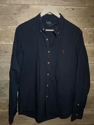 Mörkblå skjorta från Ralph Lauren - Klassisk mörkblå skjorta från Ralph Lauren med knappkrage och röd broderad logga på bröstet. Skjortan har långa ärmar, knappar framtill och är tillverkad i mjuk bomull. Perfekt för en stilren och avslappnad look.