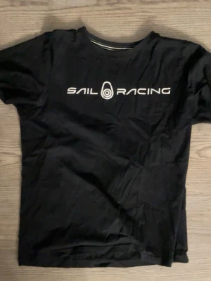 Svart Sail Racing t-shirt - Svart t-shirt från Sail Racing med vit logga och text på bröstet samt en mindre logga i nacken. Klassisk rund hals och korta ärmar. Tillverkad i mjuk bomull för en skön känsla. Perfekt för dig som gillar stilrena och sportiga plagg.