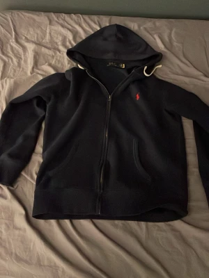 Marinblå hoodie från Polo Ralph Lauren - Mörkblå hoodie med dragkedja från Polo Ralph Lauren. Klassisk design med huva och två fickor framtill. Liten röd broderad logga på bröstet. Perfekt för en chill och stilren look. Materialet känns mjukt och bekvämt.