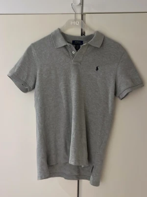 Grå pikétröja från Polo Ralph Lauren - Klassisk grå pikétröja från Polo Ralph Lauren med korta ärmar, krage och knappar framtill. Tröjan har den ikoniska broderade loggan på bröstet och är tillverkad i mjuk bomull. Perfekt för en stilren och avslappnad look storlek L i barn 14-16 år.