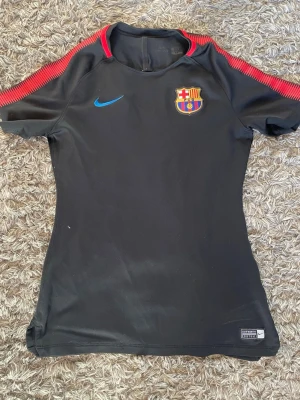 Svart FC Barcelona träningströja Nike - Svart träningströja från Nike med FC Barcelona-emblem på bröstet och blå Nike-logga. Röda detaljer längs axlarna. Perfekt för fotbollsträning eller sport. Materialet är troligtvis polyester för bra andningsförmåga.