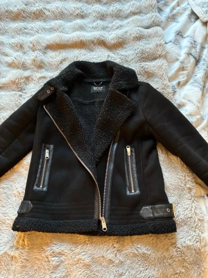 Svart mochajacka från ONLY - Säljer en svart mochajacka från ONLY med sneddragna silverfärgade dragkedjor och två fickor framtill. Jackan har en bred krage med mjukt foder och detaljer i skinn vid fickor och nederkant. Perfekt för dig som vill ha en cool och stilren look.