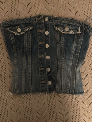 Denim korsettopp med knappar - Säljer en snygg denim korsettopp med silverfärgade knappar framtill och två dekorativa bröstfickor. Toppen har råa kanter nertill och smock baktill för skön passform. Perfekt för dig som vill ha en edgy och trendig look.