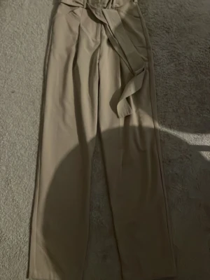 Beige kostymbyxa med knytskärp - Snygga beige kostymbyxor från Cubus med hög midja och tillhörande knytskärp. Byxorna har raka ben och bälteshällor, perfekta för en clean och stilren look. Materialet känns mjukt och följsamt, vilket gör dem bekväma att bära hela dagen.