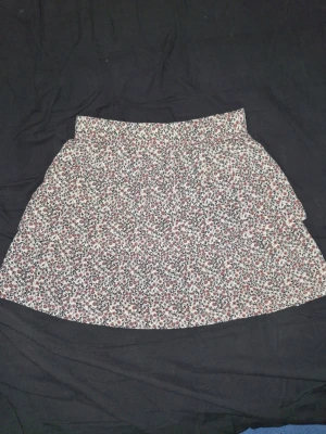 Blommig kortkjol - Supersöt kortkjol med småblommigt mönster i rosa och svart på vit botten. Kjolen har en lätt utställd form och är gjord i mjuk bomull, perfekt för varma dagar. Elastisk midja för skön passform.