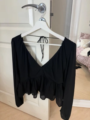 Svart blus med volang och v-ringning - Svart blus med djup v-ringning, volang nertill och långa ärmar. Blusen har knyt i nacken 🖤