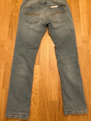 Ljusblå raka jeans från Nudie - Säljer ett par ljusblå raka jeans från Nudie Jeans med klassisk femficksmodell. Dem är oanvända och i riktigt bra skick. Jeansen har subtila slitningar framtill och bakfickor med Nudie-logga. Tillverkade i mjuk denim med en avslappnad passform. Hör av vid frågor och funderingar!