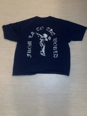 Svart t-shirt med tryck från YLA - Cool mörkblå t-shirt från Young LA med stort grafiskt tryck på ryggen där det står 'FROM LA TO THE WORLD' och en figur i mitten. På bröstet finns en liten vit Young LA-logga. T-shirten har rund hals och korta ärmar, perfekt för gymmet.