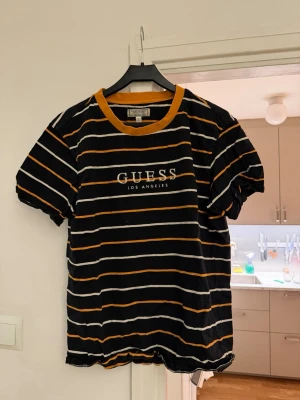 Randig svart t-shirt från Guess - Säljer en svart t-shirt från Guess med vita och orangea ränder samt orange krage. Tryck med 'GUESS LOS ANGELES' på bröstet. Skön passform och perfekt för en avslappnad stil.