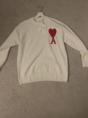 Vit stickad tröja från Ami - Vit stickad tröja från Ami Alexandre Mattiussi med röd hjärtlogga och bokstaven A på bröstet. Tröjan har rund halsringning och ribbstickade muddar. Perfekt för dig som gillar stilrena och trendiga plagg.