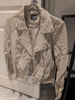 Beige skinnjacka från Livelo - Snygg beige skinnjacka från Livelo med coola detaljer som snedställd dragkedja, quiltade axelpartier och flera fickor med dragkedja. Jackan har en klassisk bikerstil och är perfekt för dig som vill ha en stilren men edgy look. Legat nerpackad därav skrynklig