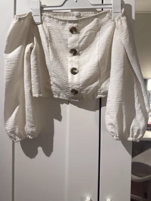 Vit offshoulder blus med knappar - Trendig vit offshoulder blus med ballongärmar och snygga knappar framtill. Toppen har en croppad passform och resår i ryggen för extra komfort. Perfekt för dig som vill ha en stilren och luftig topp till sommaren.