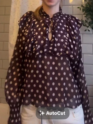 Prickig volangblus med knytning - Snygg blus i transparent material med mörklila botten och vita prickar. Blusen har volangdetaljer över axlar och bröst samt knytning vid halsen. Långa ärmar med lätt puff och avslappnad passform. Perfekt för dig som gillar trendiga mönster och detaljer.