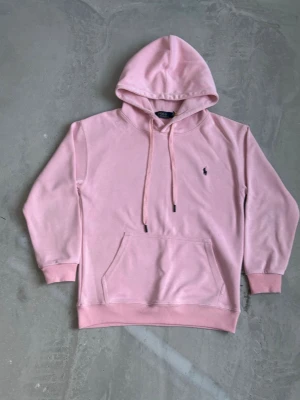 Ljusrosa hoodie från Polo Ralph Lauren - Snygg ljusrosa hoodie från Polo Ralph Lauren med klassisk broderad logga på bröstet. Hoodien har huva med dragsko, magficka och ribbade muddar. Perfekt för en chill och stilren look.