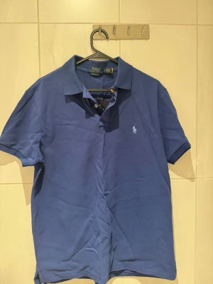 Blå pikétröja från Ralph Lauren - Klassisk blå pikétröja från Ralph Lauren med korta ärmar och broderad logga på bröstet. Tröjan har krage och knäppning framtill. Perfekt för en stilren och avslappnad look. Slimfit strl M 