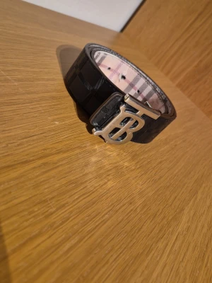 Svart rutigt Burberry bälte - Snyggt svart bälte från Burberry med klassiskt rutigt mönster och en stor silverfärgad B-logga som spänne. Insidan har den ikoniska Burberry-rutan i beige, rött och svart men lite skadat. Perfekt accessoar för att lyfta din outfit.