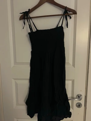 Svart långklänning från Ellos - Svart långklänning från Ellos med smock upptill och knytbara axelband. Klänningen har en luftig och böljande kjol med volangdetaljer, perfekt för varma dagar. Materialet känns mjukt och lätt, troligen bomull. Enkel och stilren design.