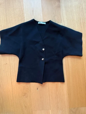 Svart kortärmad blus med knappar - Stilren svart blus från Zara med v-ringning och två dekorativa guldknappar framtill. Modellen har korta, vida ärmar och rak passform. Använd 1 gång.