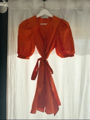 Orange kort klänning med puffärm - Trendig orange kortklänning med puffärmar och knytband i midjan. Klänningen har knappar framtill och en luftig passform som ger en cool och modern vibe. Perfekt för dig som vill sticka ut med färg och volym i garderoben.