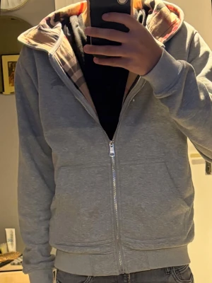 Grå Burberry hoodie  - Snygg grå hoodie från Burberry med dragkedja och klassiskt rutigt foder i huvan. Mjuk bomullskänsla, två fickor fram och ribbade muddar. Perfekt för en chill och stilren look.