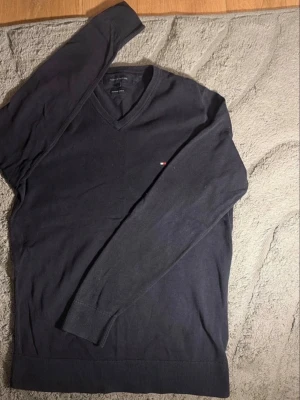 Mörkblå V-ringad tröja Tommy Hilfiger - Klassisk mörkblå V-ringad tröja från Tommy Hilfiger med diskret broderad logga på bröstet. Tröjan har långa ärmar och ribbade muddar vid ärmslut och nederkant. Perfekt för en stilren och enkel look.