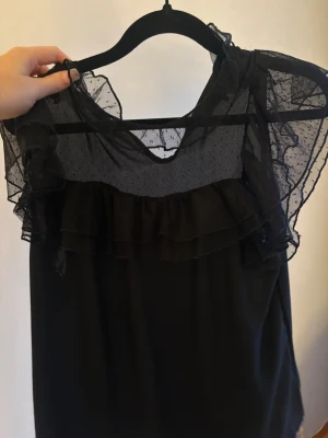 Svart blus med volang och mesh - Snygg svart blus med transparent mesh upptill och volangdetaljer runt axlar och halsringning. Blusen har prickigt mönster i meshen och är ärmlös, vilket ger en trendig och luftig känsla. Perfekt att styla med jeans eller kjol för en cool look.