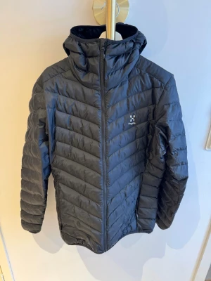 Svart dunjacka från Haglöfs med huva - Snygg svart dunjacka från Haglöfs med huva och dragkedja framtill. Västen har ett quiltat mönster och logga på bröstet. Perfekt för lager-på-lager och håller dig varm under kyliga dagar. Tillverkad i lätt och smidigt material. Litet hål i ryggen jag inte ens själv märkt. Långt ner på ena sidan. Inget som helst man märker av. I övrigt helt i nyskick, endast provad.