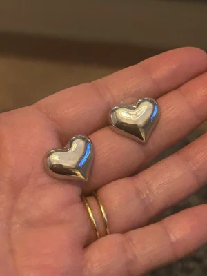 Silvriga hjärtformade örhängen - Säljer ett par stilrena örhängen i blankt silverfärgat stål material, formade som hjärtan. De har en modern och chunky look som verkligen syns. Perfekta för dig som gillar statement-smycken och vill addera lite extra till din outfit.