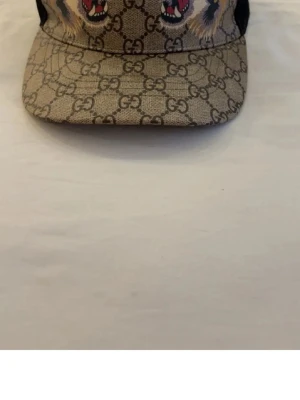 Gucci keps med GG-mönster och tigrar - Snygg keps från Gucci med klassiskt GG-monogram i beige och brunt, dekorerad med broderade tigerhuvuden framtill. Baksidan har svart mesh och justerbart band med gröna och röda ränder. Perfekt för dig som vill sticka ut med lyxig streetstyle.