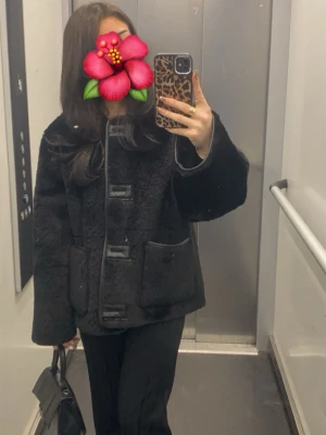 Svart pälsjacka med fickor - Säljer en svart pälsjacka med breda ärmar och stora fickor framtill. Jackan har dekorativa knäppningar och en boxig, avslappnad passform. Perfekt för att hålla sig varm under kalla dagar. Materialet är mjukt och fluffigt.