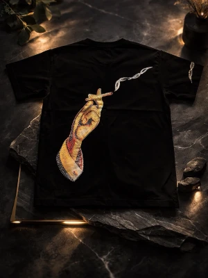 Svart Amiri t-shirt med tryck - Svart t-shirt från Amiri med korta ärmar. Framsidan har Amiri-loggan i glittriga stenar och ena ärmen har en liknande detalj. Baksidan har ett stort tryck av en hand som håller en cigarett med rök i stenar. Snygg och unik design i bomull.