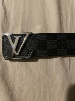 rutigt svart bälte - Snyggt svart bälte med klassiskt rutigt mönster och stor logga i silver på spännet. Bältet är gjort i skinn och har detaljer i beige på insidan. Perfekt accessoar för att lyfta din outfit.