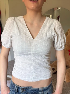 Vit v-ringad topp med puffärm - Snygg vit topp med v-ringning både fram och bak samt puffiga korta ärmar. Toppen är lätt croppad och har en smickrande passform. Tillverkad i ett tunt, strukturerat bomullstyg som känns luftigt och fräscht. Perfekt till jeans eller kjol för en trendig look. Har en liten fläck men den går bort i tvätten