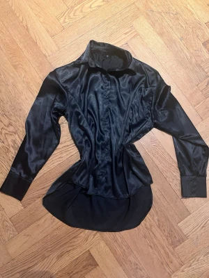 Svart satinliknande skjorta - Snygg svart skjorta i satinliknande material med lång ärm och klassisk krage. Skjortan har knappar framtill och en figurnära passform som ger en modern siluett. Perfekt för dig som vill ha en stilren och trendig look.