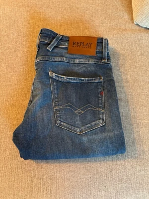 Blå jeans från Replay - Snygga blå jeans från Replay med coola slitningar. Jeansen har en skön passform (ANBASS) och detaljer som läderpatch med logga bak samt broderad bakficka. Perfekta till vardagslooken, skicket är 10/10 inga defekter som helt nya, och har haft dom över 1 år och dom är riktigt fina