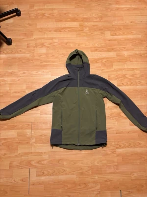 Grön och mörkgrå vindjacka Haglöfs - Snygg vindjacka från Haglöfs i olivgrönt och mörkgrått. Jackan har huva, hel dragkedja och två sidofickor med dragkedja. Perfekt för blåsiga dagar och outdoor-aktiviteter. Materialet är lätt och smidigt, med logga på bröstet.
