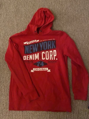 Röd hoodie New York Denim Corp - Snygg röd hoodie från Denim Corp med tryck i blått, vitt och svart på bröstet där det står 'New York Denim Corp'. Hoodien har huva med dragsko och långa ärmar. Perfekt för en avslappnad streetwear-look.