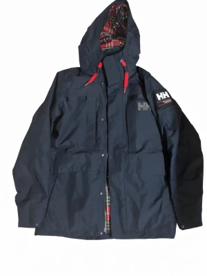 Mörkblå  Helly Hansen jacka  - Säljer en riktigt schyst Mörkblå regnjacka från Helly Hansen med huva och röda detaljer på snörena. Jackan har flera fickor framtill, HH-logga på bröstet och ärmen samt rutigt innerfoder. Perfekt för regniga dagar och outdoor-äventyr. Pälsen förekommer som originalet sitter på jackan, den kan man ta av och på om man vill. Priset kan diskuteras. Nypris: ~2150