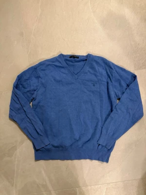Blå stickad tröja från Gant - Snygg blå stickad tröja från Gant med V-ringning och diskret logga broderad på bröstet. Tröjan har långa ärmar och ribbade muddar vid ärmslut och nederkant. Perfekt att bära över en skjorta för en clean look.