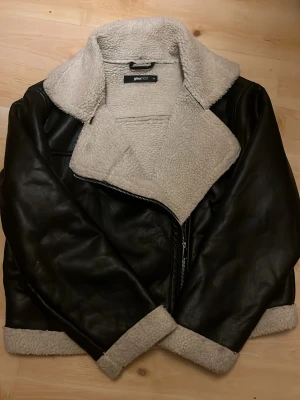 Svart pälsjacka från Gina Tricot - Snygg svart pälsjacka från Gina Tricot med bred krage och mysigt ljust teddyfoder. Jackan har dragkedja och detaljer i samma teddy vid ärmslut och nederkant. Perfekt för kalla dagar och ger en cool look.