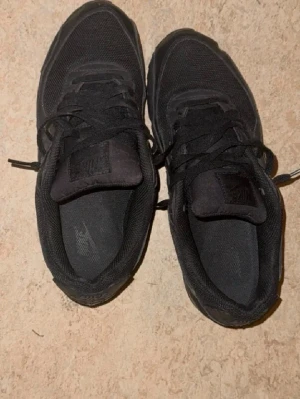 Svarta Nike Air Max 90 sneakers - Säljer ett par helsvarta Nike Air Max 90 sneakers med klassisk chunky sula och synlig Air-enhet. Skorna har ovandel i mesh och läder, samt svarta skosnören och diskret Nike-logga på sidan och plösen. Perfekta för dig som gillar en clean och sportig look.