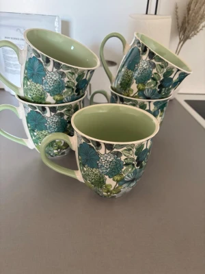 Blommiga koppar med grön insida - Fem  koppar med blommigt mönster i grönt, blått och vitt. Insidan är ljusgrön och kopparna har rund form med bekvämt handtag. Perfekta för te eller kaffe och ger en färgglad touch till köket. Materialet är porslin.
